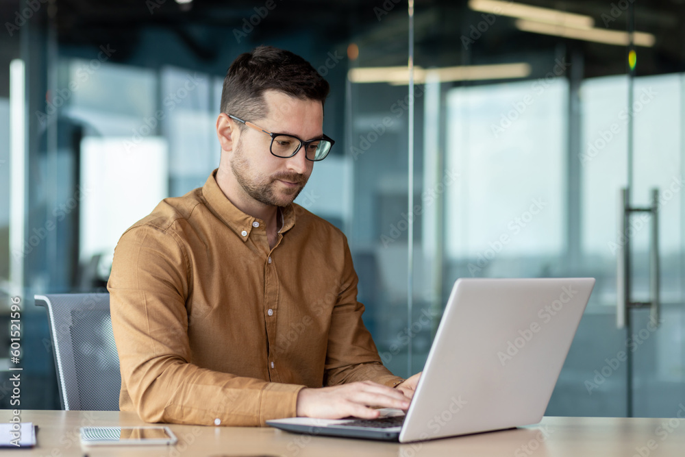 Portrait of a young male programmer in a brown shirt and glasses who is concentrating on working on a laptop while sitting at a desk in the office Portrait of a young male programmer in a brown shirt and glasses who is concentrating on working on a laptop while sitting at a desk in the office