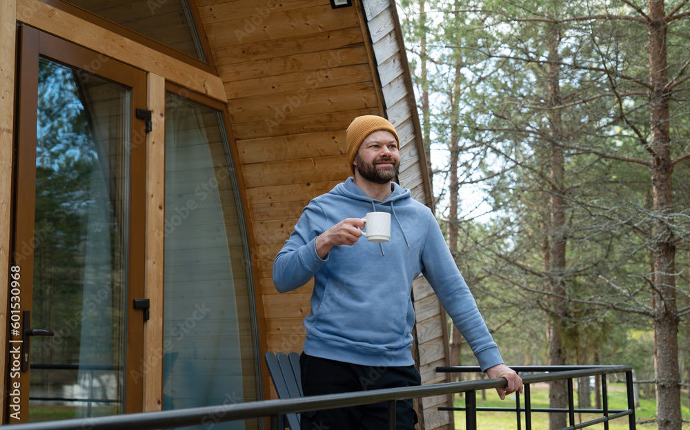 Handsome bearded man drinking hot tea in front of a modern wooden house in the forest