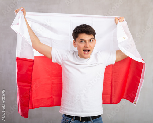 Middle-aged positive man in casual clothes holds unfurled flag of Poland in hands raised above head against gray wall, studio shot. Support, national identification photo