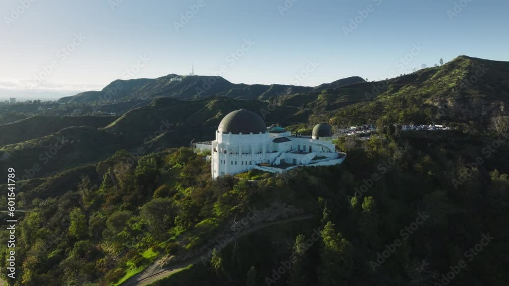 Los Angeles city California USA March 2023. Flying around dome architecture Griffith Observatory, Hollywood sign on bright green hill at sunset. Aerial scenic landmark buildings in green spring park