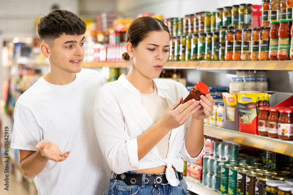 Focused interested young girl reading label on glass jar with sauce while shopping in supermarket with boyfriend