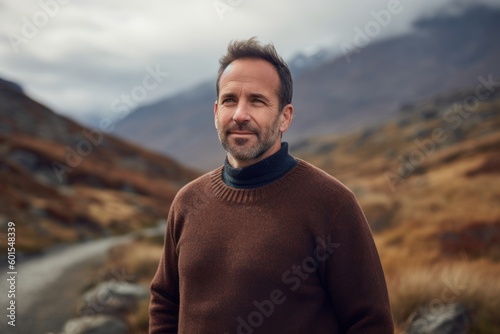 Photography in the style of pensive portraiture of a grinning man in his 40s wearing a cozy sweater against a mountain landscape background. Generative AI