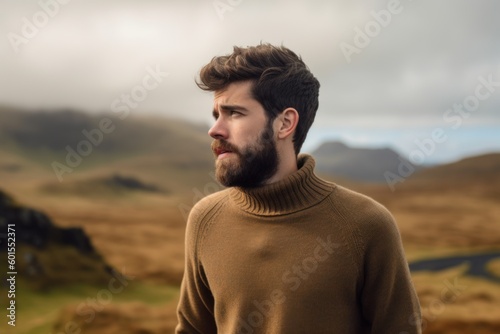 Portrait of a handsome bearded man with long hair and beard, wearing brown sweater, standing against scenic landscape of Scotland.