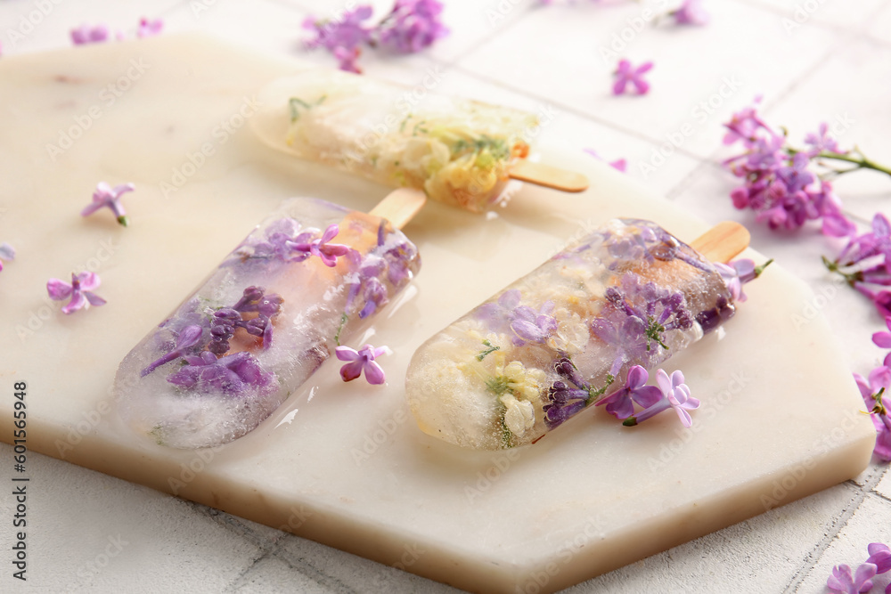 Board with beautiful floral popsicles and lilac flowers on light tile background Board with beautiful floral popsicles and lilac flowers on light tile background