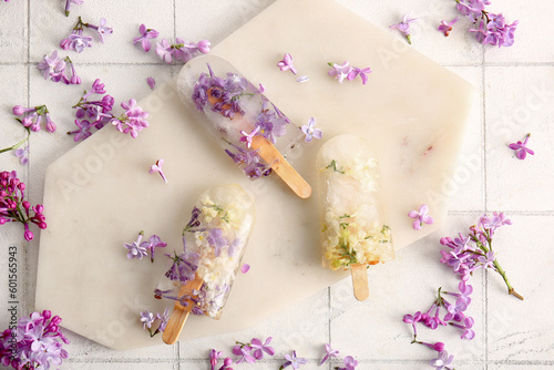 Board with beautiful floral popsicles and lilac flowers on light tile background
