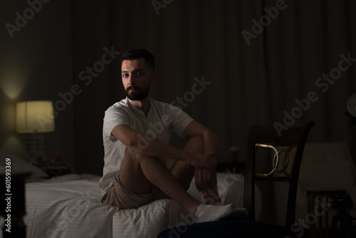 A young man, seated on the edge of a bed in a dimly lit room, has a serious expression. Dramatic lighting on his face highlights his handsome features and dark beard.