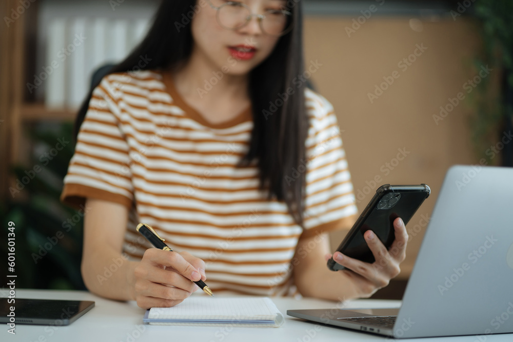 Woman hand working at a smartphone, computer and writing on a notepad with a pen in the home office. Woman hand working at a smartphone, computer and writing on a notepad with a pen in the home office.