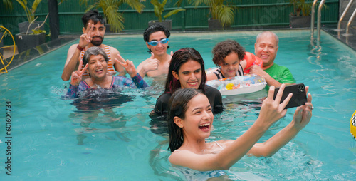 Happy Indian family fun enjoy outdoor picnic teen adult girl take selfie show thums up with parents using mobile smartphone at blue water park cute child relax in colorful floating ring tube at resort