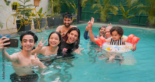 Happy Indian family fun enjoy outdoor picnic teen adult boy take selfie show thums up with parents using mobile smartphone at blue water park cute child relax in colorful floating ring tube at resort photo