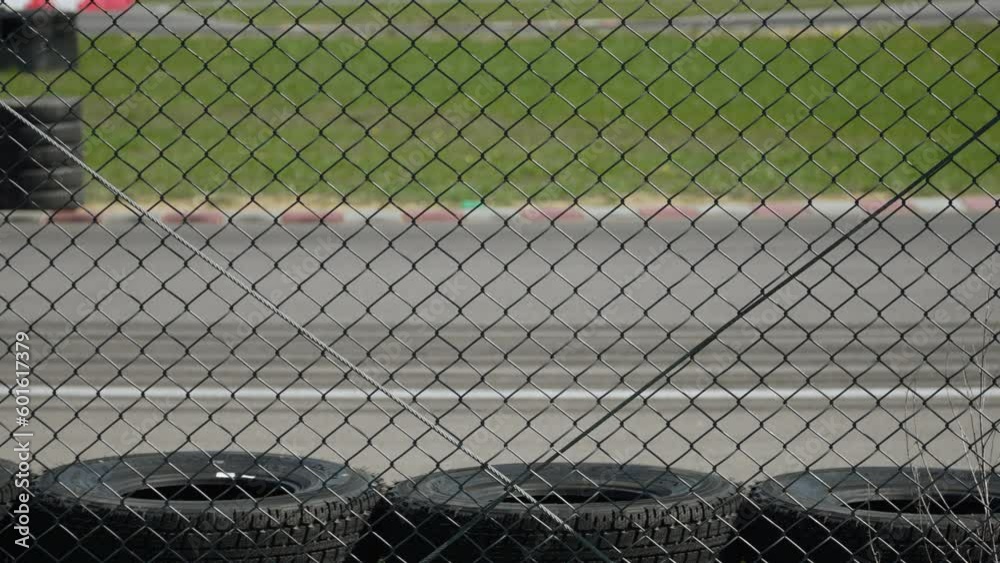 Metal mesh fencing and collision energy absorption zone from car tires on race track. Through corner, near red white curb, blue sports hatchback passes at sunny day. Metal mesh fencing and collision energy absorption zone from car tires on race track. Through corner, near red white curb, blue sports hatchback passes at sunny day.