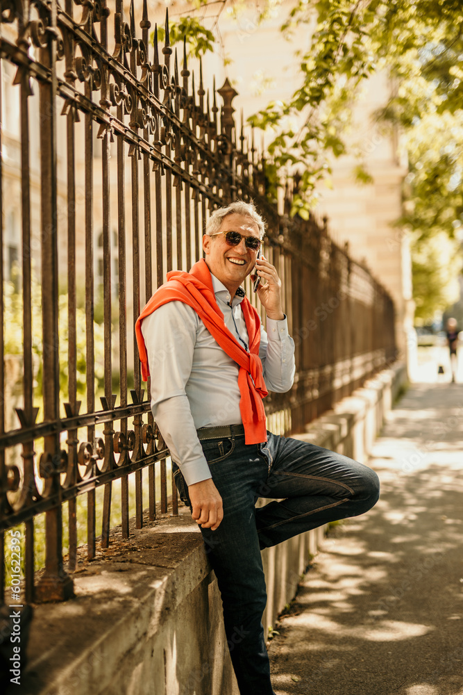 Fototapeta premium Senior man standing leaning on the fence, using a phone and relaxing after a city shopping.