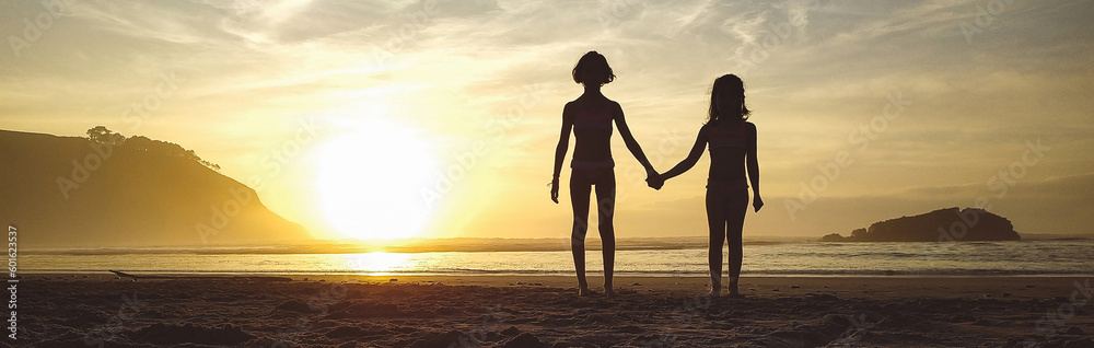 Silhouettes of two unrecognizable little girls holding her hands in the beach at sunset