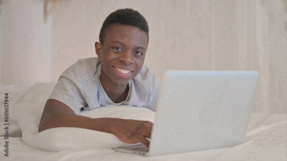 Smiling Young African Man Looking in Camera while using Laptop Lying on Stomach Smiling Young African Man Looking in Camera while using Laptop Lying on Stomach