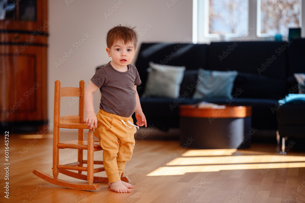 Little toddler boy sitting on wooden rocking chair in living room with dark sofa Little toddler boy sitting on wooden rocking chair in living room with dark sofa