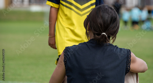 Mom supporting her son playing football in a school tournament on a sideline with a sunny day. Sport, outdoor active, lifestyle, happy family and soccer mom and soccer dad concepts.