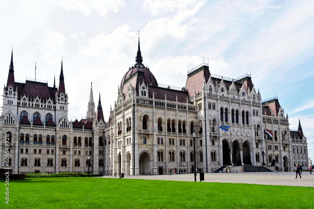 The Hungarian Parliament in Budapest. large main dome and smaller towers. arched Gothic windows. fresh bright green lawn. visitors' silhouettes in a distance. famous popular landmark. open square.