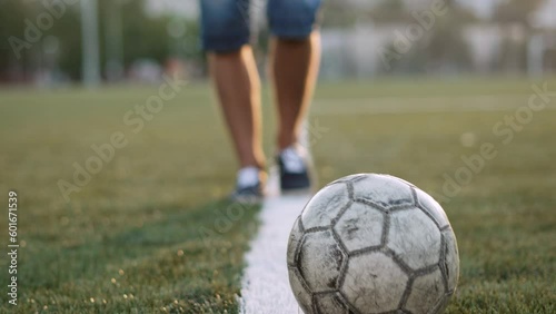 Unrecognizable football player passes old shabby soccer ball, close-up. Ball rolls across football field along white line of penalty area at sunset. Dynamic camera follows rolling sports equipment. photo