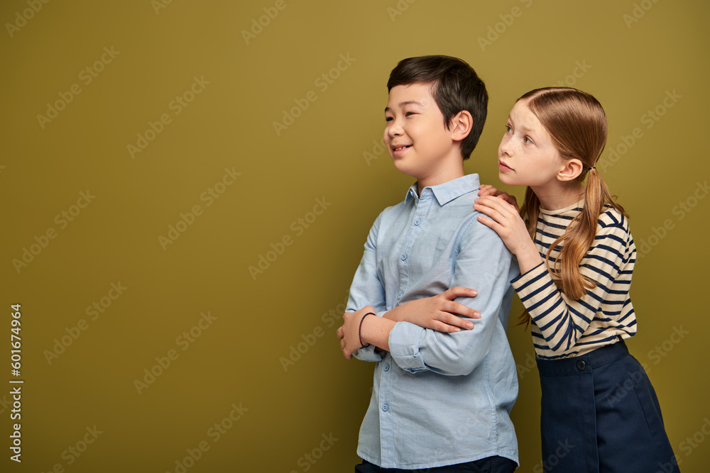 Redhead girl in striped blouse hugging smiling asian friend crossing and and looking away during child protection day celebration on khaki background with copy space Redhead girl in striped blouse hugging smiling asian friend crossing and and looking away during child protection day celebration on khaki background with copy space