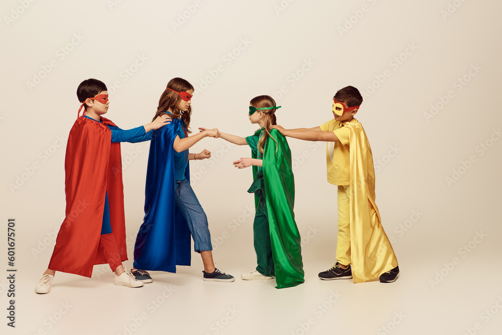 side view of interracial kids in colorful superhero costumes with masks and cloaks fighting with each other on grey background in studio, International children's day concept