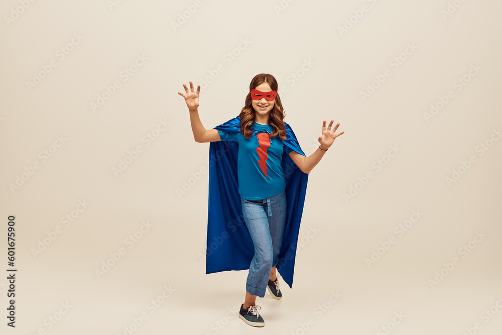 joyful girl in superhero costume with blue cloak and red mask on face, showing hand gesture, standing in denim jeans and t-shirt while celebrating Child protection day holiday on grey background joyful girl in superhero costume with blue cloak and red mask on face, showing hand gesture, standing in denim jeans and t-shirt while celebrating Child protection day holiday on grey background