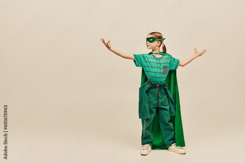 powerful girl in superhero costume with green cloak and mask on face standing with outstretched hands while showing strength and celebrating International children's day on grey background