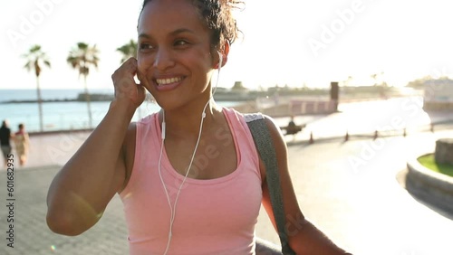 Smiling young African woman in sportswear listening to music on earphones while crossing the street carrying a yoga mat