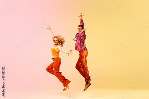 Dynamic image of beautiful young couple, man and woman in stylish vintage clothes dancing against gradient pink yellow background. Concept of retro style, dance, fashion, art, hobby, music, 70s
