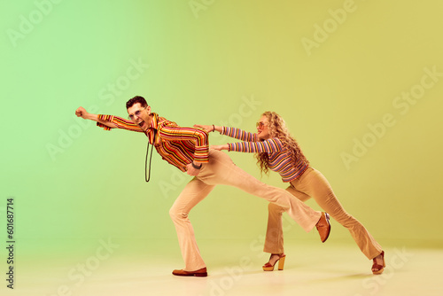 Expressive, talented young couple, man and woman in stylish vintage costumes dancing against gradient green yellow background. Concept of retro style, disco dance, fashion, art, hobby, music, 70s