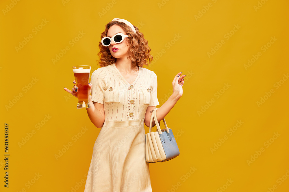 Young beautiful girl wearing stylish retro clothes with sunglasses holding glass of beer over yellow background. Vintage fashionista - obrazy, fototapety, plakaty 