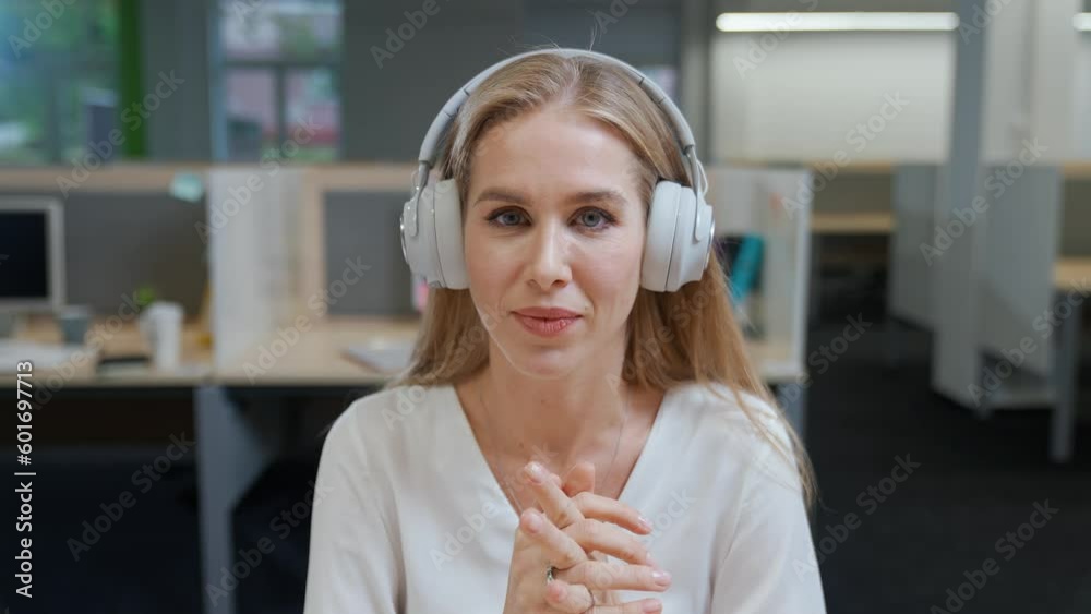 Portrait of a cheerful woman designer against the background of international office space, young woman in headphones communicates by video call, talks looking at the camera, template.