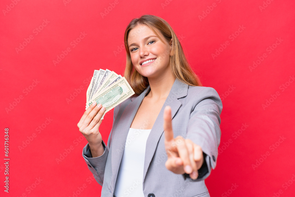 Young business caucasian woman holding money isolated on red background showing and lifting a finger Young business caucasian woman holding money isolated on red background showing and lifting a finger