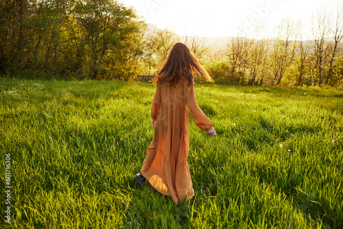 an attractive, slender, red-haired woman walks through a field during sunset, in a long orange dress enjoying unity with nature and relaxation holding the edge of her clothes