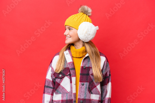 Young beautiful woman wearing winter muffs isolated on red background looking side
