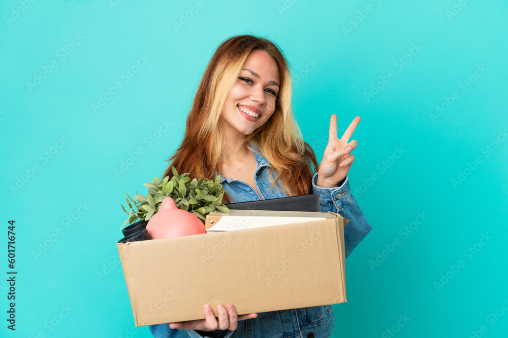 Naklejka premium Teenager blonde girl making a move while picking up a box full of things smiling and showing victory sign