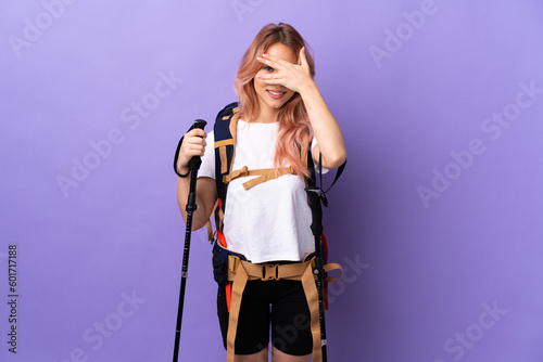 Teenager girl with backpack and trekking poles over isolated purple background covering eyes by hands and smiling
