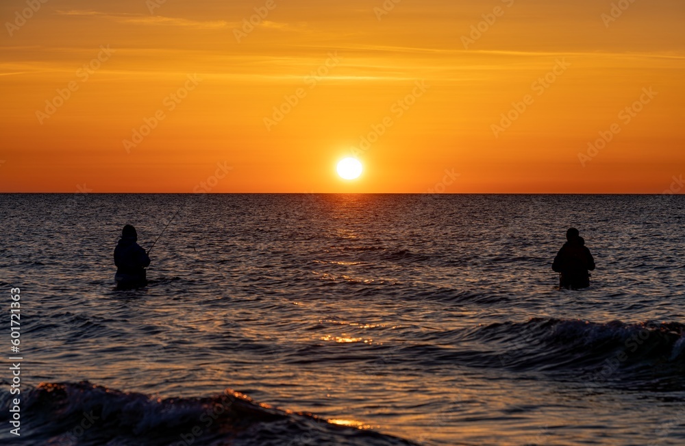Die Silhouetten von zwei  Anglern in wasserdichten Hosen angelen vor einem traumhaften orangen Sonnenuntergang  in der Ostsee. Sie stehen mitten im Wasser der Ostsee.