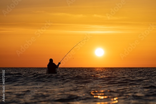 Ein Angler in wasserdichten Hosen angelt vor einem traumhaften orangen Sonnenuntergang in der Ostsee. Er steht mitten im Wasser der Ostsee.