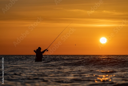 Silhouette eines Angler in wasserdichten Hosen welcher vor einem traumhaften orangen Sonnenuntergang in der Ostsee angelt / fischt . Er steht mitten im Wasser der Ostsee.