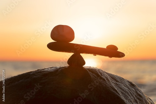 Schieflage / Dysbalance der Kräfte - natürliche Wage aus Steinen ( im Ungleichgewicht) am Meer zum orangenen Sonnenuntergang - Symbolfoto	
