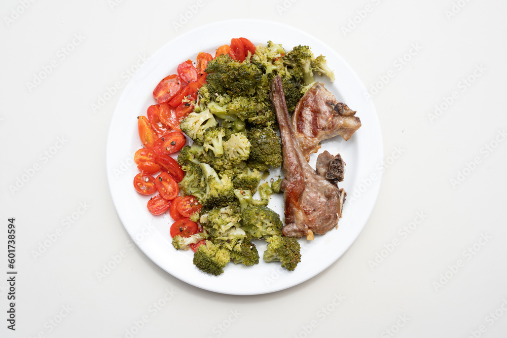 White plate on white background with sunlight, steamed broquil, cherry tomatoes and two grilled lamb ribs. Nutritious, colourful and varied dish.