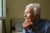 Portrait of a senior female sitting on the bed and leaning on a cane. Elderly woman resting her head on a handle of a walking cane. Close up, copy space for text, background.