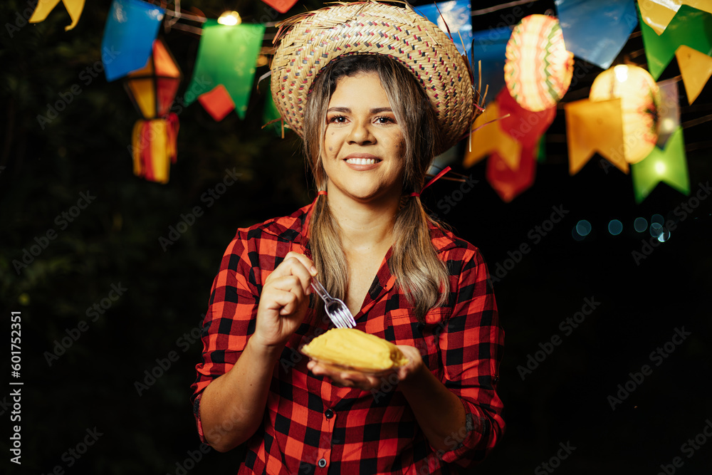 Woman eating Pamonha, a traditional corn-based dessert, at the Festa Junina (June Festival) embracing the flavors and traditions of Brazil's June celebration. - obrazy, fototapety, plakaty 