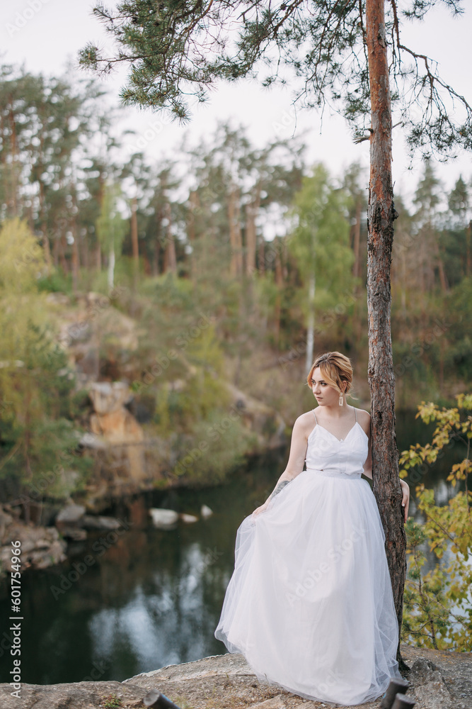 Young bride stands on rock against background of lake, forest and canyon.