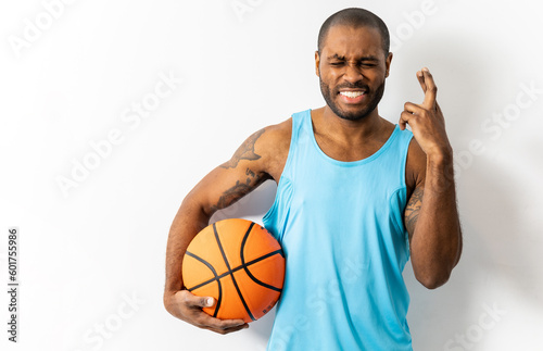Dark-skinned adult male poses with a basketball on a white background wearing a tank top. The boy is crossing with the fingers of his hand, waiting for the result, imagining the luck.