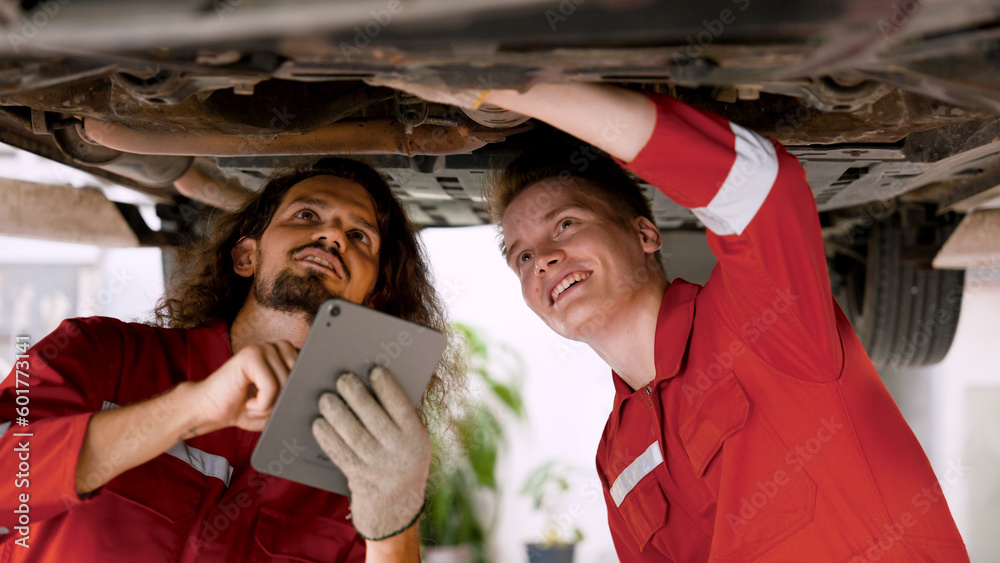 two men who are mechanics, working in large garage, two mechanics checking undercarriage car raised high see lower part clearly, holding car check book talk each other before fixing car. two men who are mechanics, working in large garage, two mechanics checking undercarriage car raised high see lower part clearly, holding car check book talk each other before fixing car.