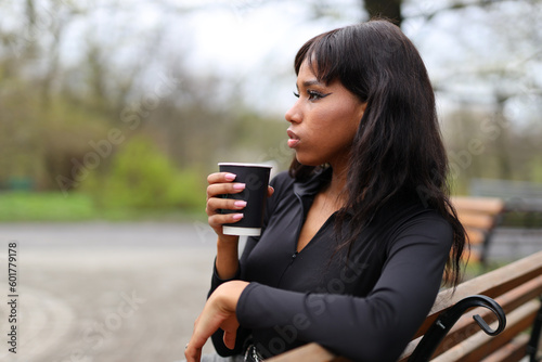 Young African girl is relaxing with takeaway coffee on a bench in the park.