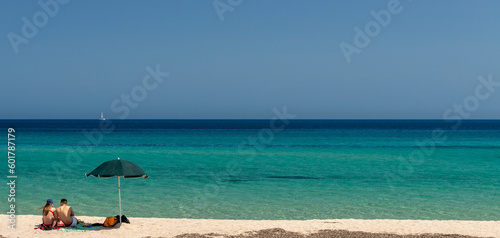 Couple on the beach in front of a beautiful and turquoise sea and blue sky with no clouds. Young people in romantic leisure at a paradise destination. Mediterranean sea, Sardinia, Italy.