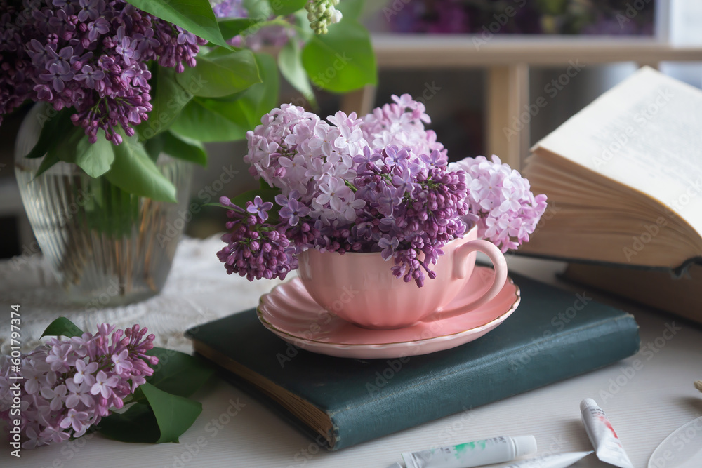 Romantic background with a bouquet of lilacs in a cup, lilac flowers and an open book, scattered paints on a white table. Romantic background with a bouquet of lilacs in a cup, lilac flowers and an open book, scattered paints on a white table.