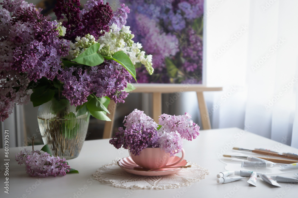 Romantic background with a bouquet of lilacs in a cup, lilac flowers and an open book, scattered paints on a white table.