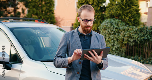 Attractive and smart adult business man holding in hands tablet, standing near car in parking lot in residential area . Outdoors. Busy lifestyle